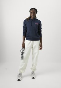 Mornarska dukserica s prednjim džepom i ružičastim logom, u kombinaciji s bijelim trenirkama i sivim tenisicama, drži užad za preskakanje. Glatke teksture kroz cijelu odjeću.