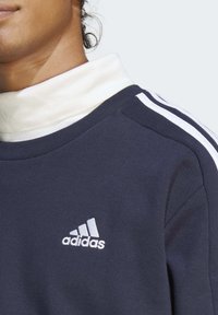 Mornarska dukserica s bijelim visokim ovratnikom, tri bijele pruge na ramenima i srebrnim izveznim Adidas logotipom.