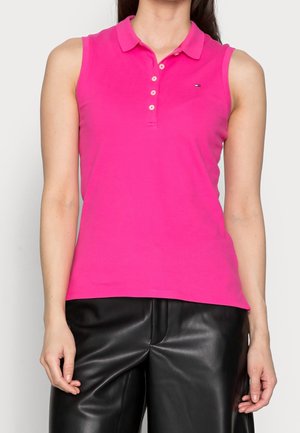 Vrouw draagt een mouwloze felroze poloshirt met knopen, gecombineerd met een zwarte leren broek, tegen een effen lichte achtergrond.