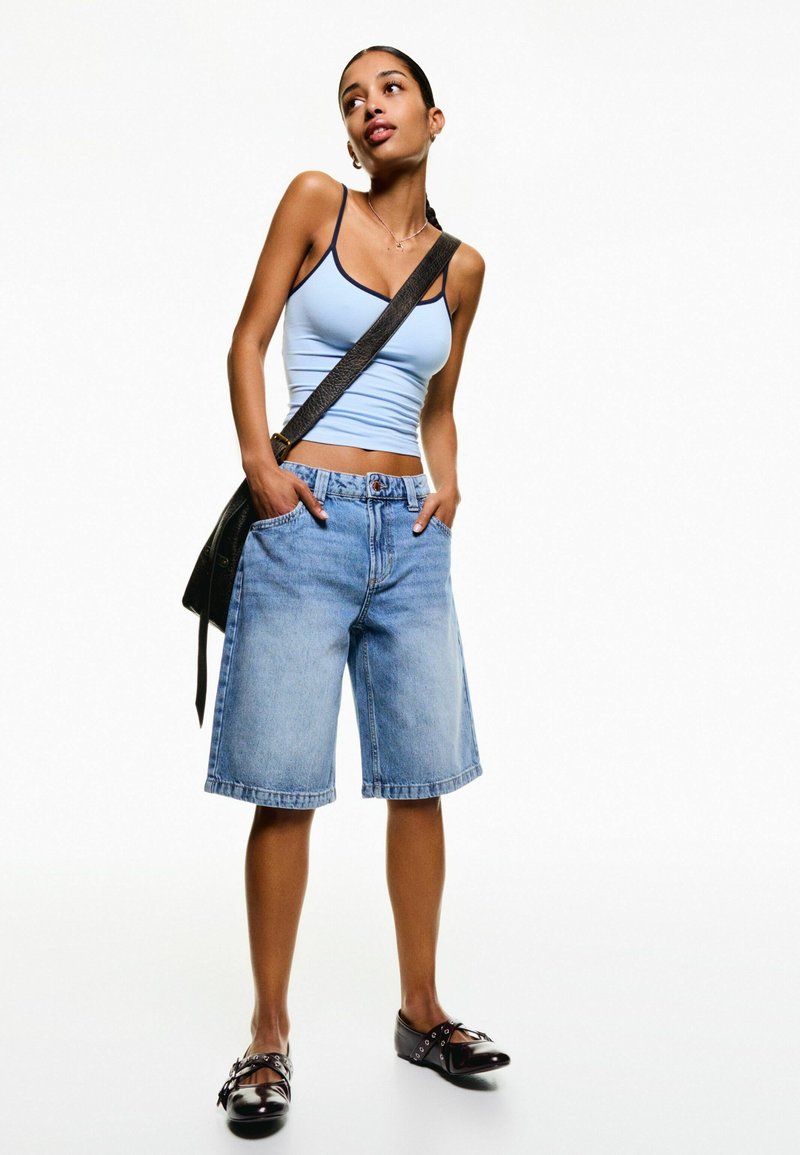 Débardeur bleu clair, shorts en denim taille haute, et chaussures noires avec quincaillerie décorative. Le modèle tient un sac à bandoulière noir, posé contre un fond blanc.