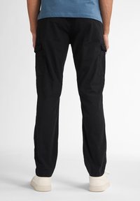 Zwarte cargos met zijzakken, elastische boorden en een ontspannen pasvorm. Gecombineerd met witte sneakers en een blauwe shirt. Alleen achteraanzicht.