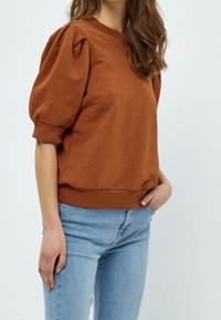 Brown sweatshirt with puffed sleeves and ribbed cuffs, paired with light blue jeans. Material appears soft and slightly textured.