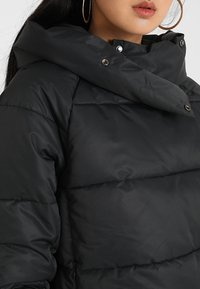 Femme portant une doudoune noire avec un col haut et des boutons-pression, montrant ses lèvres, une boucle d'oreille, et de longs cheveux foncés.