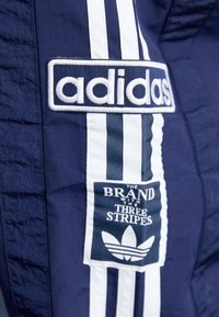 Námořnické sportovní kalhoty s bílými látkovými pruhy a vyšitým logem Adidas. Obsahují nášivku s nápisem "Značka se třemi pruhy."