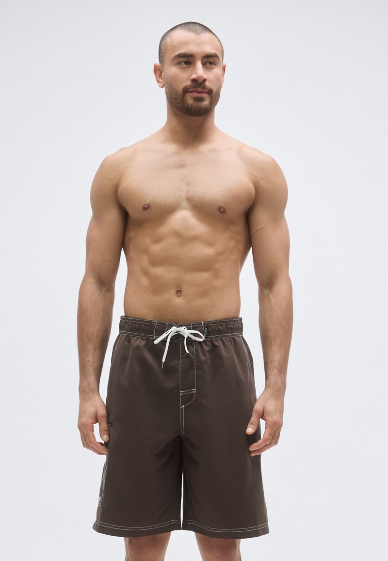 Hnedé plavkové šortky s voľným strihom, vybavené bielym sťahovacím šnúrkou a kontrastným prešívaním. Materiál pôsobí ľahko a je odolný voči vode.