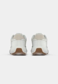 Um par de ténis brancos vistos de trás, com detalhes em bege e solas castanhas estampadas com o logótipo "cK".