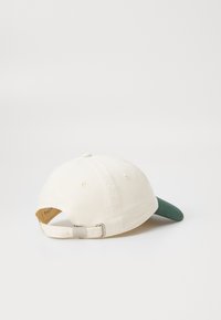 Baumwollkappe mit einem cremefarbenen Körper und einem dunkelgrünen Schirm, ausgestattet mit sechs Belüftungsösen und einem verstellbaren Riemen mit einer Metallbuckle.