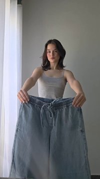 Νεαρή γυναίκα με γκρι αμάνικο τοπ κρατά μπροστά της μπλε-γκρι τζιν παντελόνι μεγάλου μεγέθους, με φόντο έναν μονόχρωμο γκρι τοίχο και μιας λευκής διαφανούς κουρτίνας.