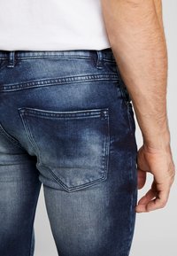 Hombre vestido con jeans de mezclilla azul desgastados y una camisa blanca, visto desde atrás con una mano relajada a su lado.