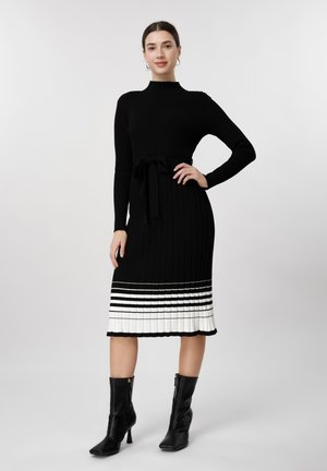 Naine musta pika varrukaga kudumkleidis, millel on valge triibuline ääris, ja mustade pahkluu-saabastega, seisab ühe käega puusal ühevärvilise tausta ees.