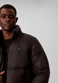 Homme portant une doudoune noire Calvin Klein par-dessus un pull noir, sur un fond uni gris clair.