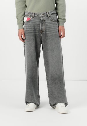 Osoba nosząca luźne szare, sprane jeansy z szerokimi nogawkami, zieloną koszulę z długim rękawem, białe trampki oraz częściowo widoczny biało-czerwony przedmiot w kieszeni.