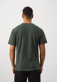 Camiseta de algodón verde oscuro con mangas cortas, cuello redondo y una textura desgastada. Presenta un corte relajado y detalles de costura sutiles.