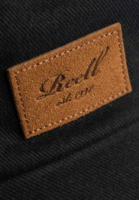 Gorra de tela negra con un parche rectangular de cuero marrón. El parche presenta texto en relieve "Reell" y "est. 1991" cosido a lo largo de los bordes.