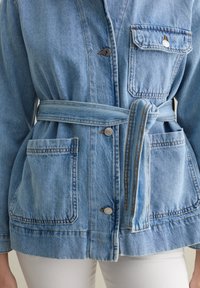 Giacca in denim azzurro chiaro con vita a cintura, due tasche frontali e chiusura a bottoni argentati. Cuciture visibili e texture morbida.