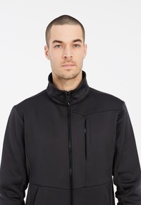 Sort atletisk jakke med høj krave, lynlås foran, brystlomme og strukturerede sidelapper. Stoffet ser glat og tætsiddende ud.