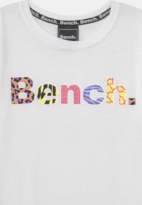Biała koszulka z nadrukiem słowa „Bench.” w wielokolorowych literach z motywami zwierzęcymi. Okrągły dekolt, miękki materiał i kołnierzyk z logo marki.