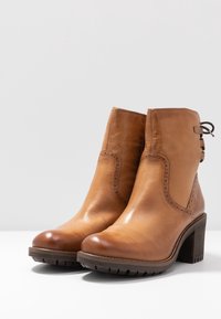 Hnědé kožené kotníkové boty s hladkým povrchem, zaoblenou špičkou, dekorativním bočním prošitím a blokovým podpatkem. Na zadní straně mají tkaničky.