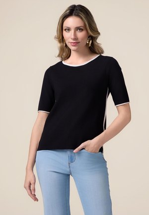 Top nero con maniche corte, caratterizzato da un collo rotondo con bordi bianchi, texture liscia e spacchi laterali. Abbinato a jeans azzurri chiari.