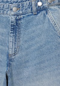 Krupan plan svijetloplavih traperica koji prikazuje prednji džep, petlju za remen i metalne gumbe s reljefnim tekstom.