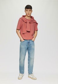 Roze shirt met korte mouwen, met een trui om de nek gedrapeerd, blauwe spijkerbroek en witte sneakers met beige accenten. Simpele, casual outfit.