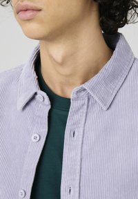 Chemise boutonnée en velours côtelé violet clair avec une texture douce, comportant un col évasé, des boutons visibles, et un t-shirt à col rond vert en dessous.
