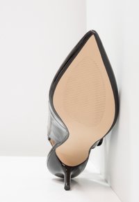 Chaussure à talon haut en cuir verni noir avec bout pointu reposant sur une surface blanche et un mur blanc en arrière-plan, semelle vers l'avant.