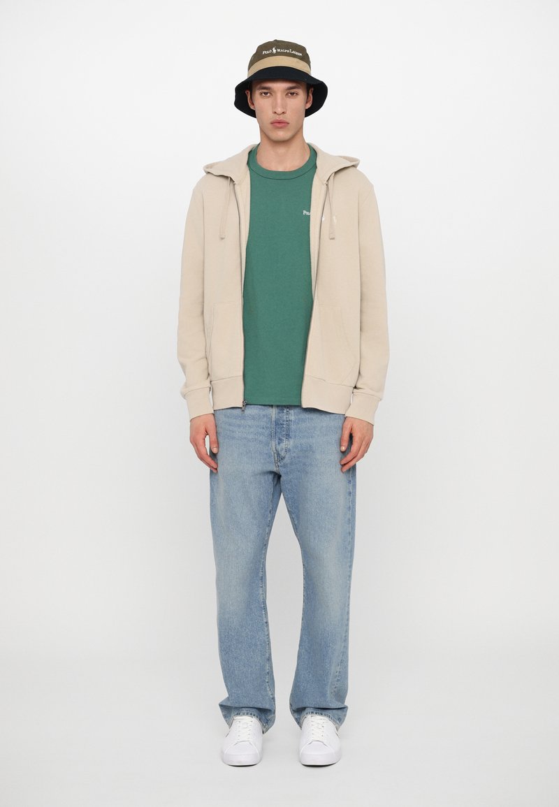 Beige zip-up hoodie over een groene t-shirt, gecombineerd met lichtblauwe jeans en witte sneakers, afgemaakt met een donkergroene bucket hat.