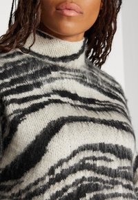 Pull en maille à col roulé crème avec un motif rayé tigre noir. Texture douce, col côtelé et coupe légèrement oversize.