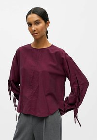 Blusa burdeos de manga larga con puños elásticos y lazos de cinta, de tejido ligero y texturizado, con escote redondo y dobladillo curvado.