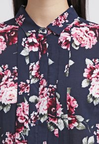 Camicia a maniche corte con bottoni, floreale, caratterizzata da uno sfondo blu navy con rose rosa e rosse. Include un colletto strutturato e pieghe verticali sul davanti.