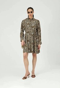 Femeie îmbrăcată cu o rochie lungă cu imprimeu leopard, ochelari de soare mici, cercei rotunzi, sandale cu toc înalt maro și purtând o poșetă mică aurie.