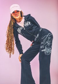 Jonge vrouw met lange vlechten draagt een zwart Ed Hardy trainingspak met panterdesign en een lichtroze pet, poseert tegen een roze achtergrond.