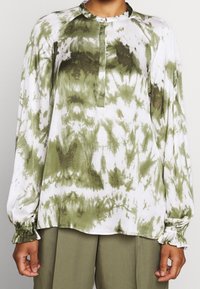 Osoba ubrana w koszulę z długim rękawem w zielono-białe tie-dye ze zgromadzonymi mankietami oraz oliwkowe spodnie, stojąca na neutralnym tle.