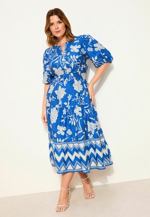 Nő térdig érő kék ruhában, fehér virág- és cikkcakkmintákkal, pöttyös ujjakkal és bokapántos szandálokban, egyszerű fal előtt állva.