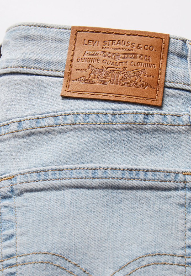 Jeans en denim bleu clair avec une pièce en cuir brun texturée portant le nom de la marque et un design en relief. Coutures visibles et détails rivetés.
