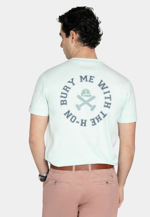 Hombre con camiseta azul claro con gráfico y texto de bates de béisbol cruzados y gorra que dice "Entiérrame con la H-on" en la espalda, pantalones rosas, cinturón marrón.