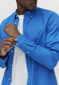 Blauwe knopjesoverhemd met een gladde textuur, uitgerust met een klassieke kraag en manchetten, gelaagd over een wit t-shirt. Subtiele knoopdetails zichtbaar.