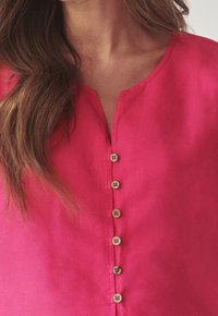 Lyserød linnedbluse med v-udskæring og otte træknapper foran. Stoffet har en blød tekstur, der subtilt reflekterer lyset.