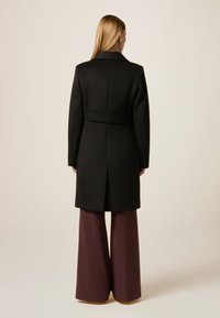 Cappotto di lana nero con silhouette su misura, apertura posteriore singola e dettaglio in vita arricciato, abbinato a pantaloni bordeaux a gamba larga.
