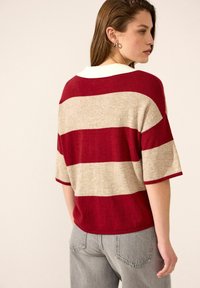 Červeno-beige pruhovaný svetr s krátkými rukávy a bílým límečkem, který má volný střih a měkkou texturu. Nosený s šedými džíny.