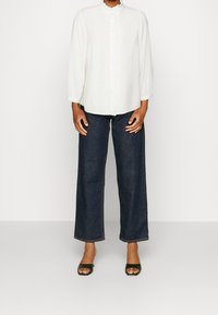 Persona che indossa una camicetta bianca a maniche lunghe, jeans ampi scuri e tacchi aperti neri in piedi contro uno sfondo neutro.