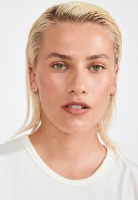 Portrét osoby s mokrými, platinově blond vlasy, oblečené v krémovém kratším tričku s žebrováním. Pleť vypadá hladce s přirozeným makeupem.