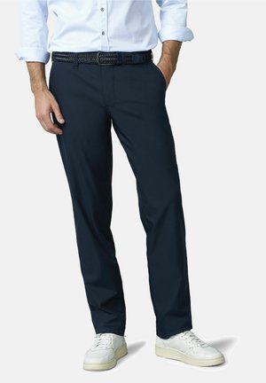 Mann trägt marineblaue Hose, weißen gewebten Gürtel, weißes Hemd mit hochgekrempelten Ärmeln und weiße Turnschuhe, steht mit einer Hand in der Tasche.