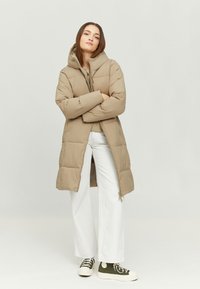 Beige puffer coat with a hood, zip closure, and a quilted design. Paired with white wide-leg pants and green high-top sneakers.