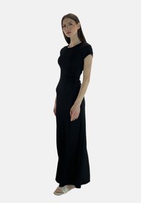 Schwarzes, figurbetontes Maxikleid mit kurzen Ärmeln und gerafftem Seitendetail, getragen mit hellfarbenen Slide-Sandalen. Glatte Textur.