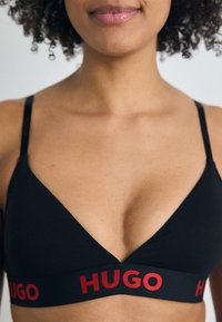 Zwarte driehoekige bralette gemaakt van zacht materiaal met dunne bandjes. Bevat een rode logoband met "HUGO" opvallend weergegeven.