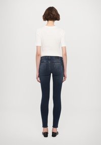 Chudé jeans v tmavom denime s hladkou štruktúrou, s polovičným pásom, zadnými vreckami a jemnú nášivkou s logom. Skombinované s bielym topom.