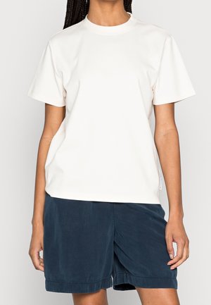 Personne portant un t-shirt blanc uni à manches courtes et un short bleu foncé à coupe décontractée, debout devant un fond uni.