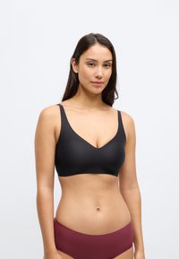 Črni brezžični modrček z izrezom v obliki črke V, gladke teksture in tankimi nastavljivimi naramnicami. Spodaj so združene z bordo brezšivnimi hlačkami.